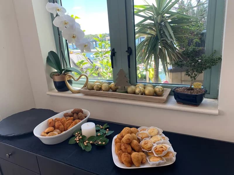Two dishes of coxinhas and empadinhas on a dark counter below a window sill of gold baubles